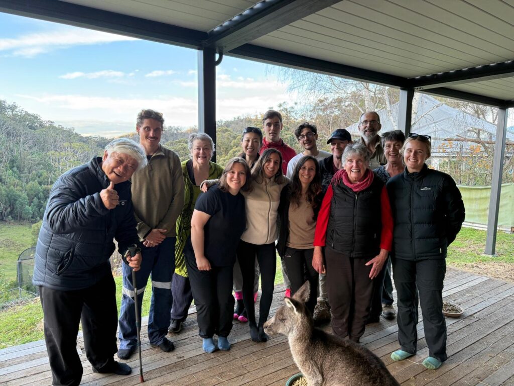 The Creating Glideways team of volunteers and hosts smiling on a verandah overlooking forested hills, with a kangaroo in the foreground.