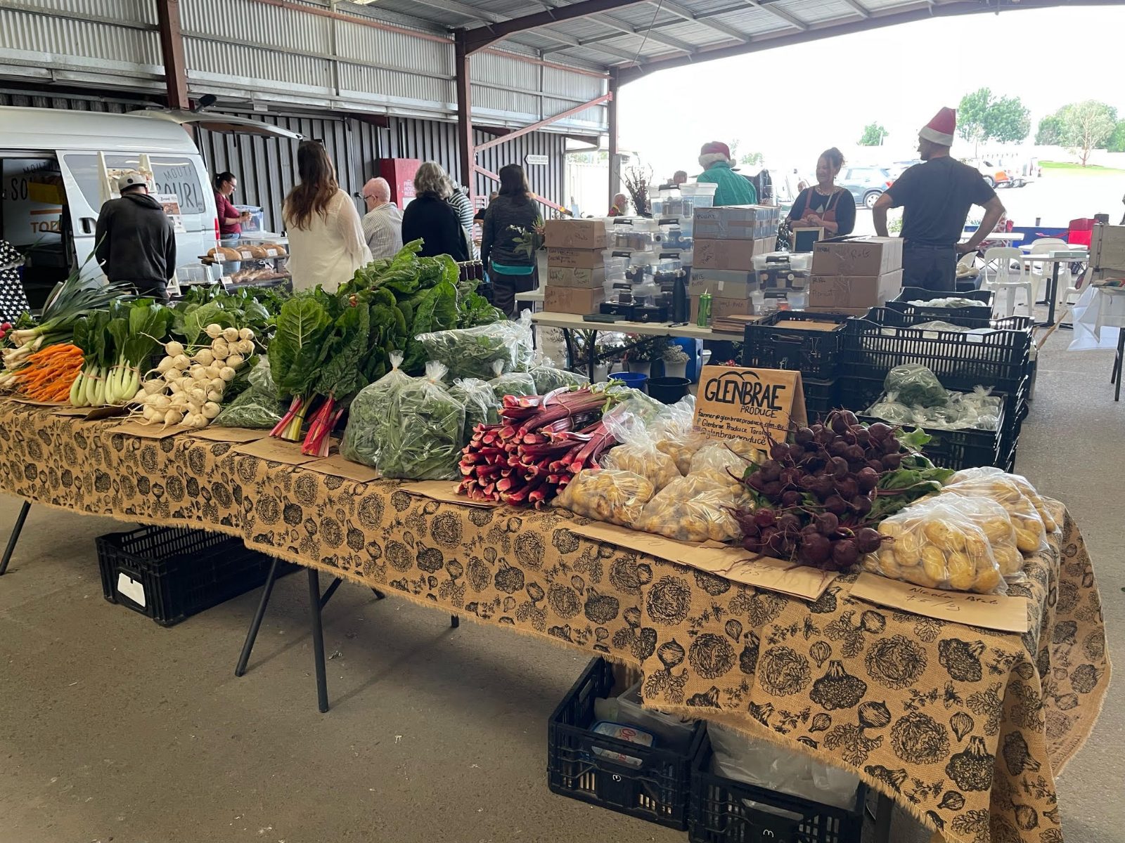 A market stall table covered with fresh vegetables, including carrots, silverbeet, rhubarb, beetroot and potatoes, with shoppers behind.