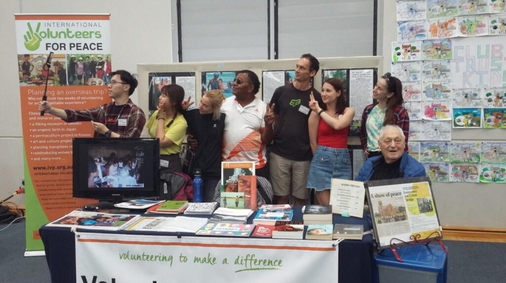 A smiling group of volunteers taking a selfie behind the IVP information table at the Goulburn Show, with an International Volunteers for Peace banner, books and photos on display.