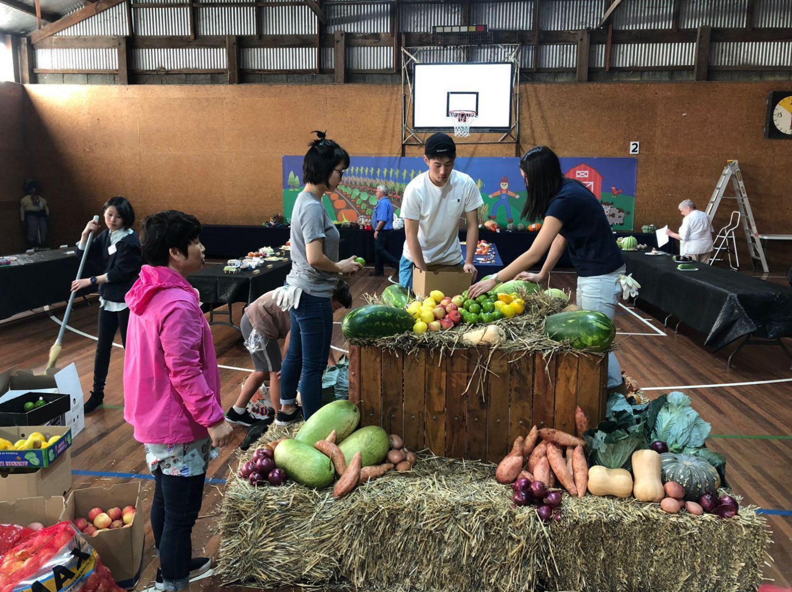 Volunteers in a show pavilion building a display of watermelons, pumpkins, sweet potatoes, apples and cabbages on hay bales and a wooden crate.