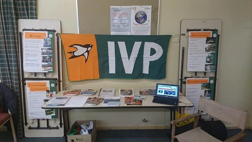 An IVP information stall: an orange and green IVP flag with a dove, hung above a table of leaflets and a laptop, with display boards about workcamps on either side.