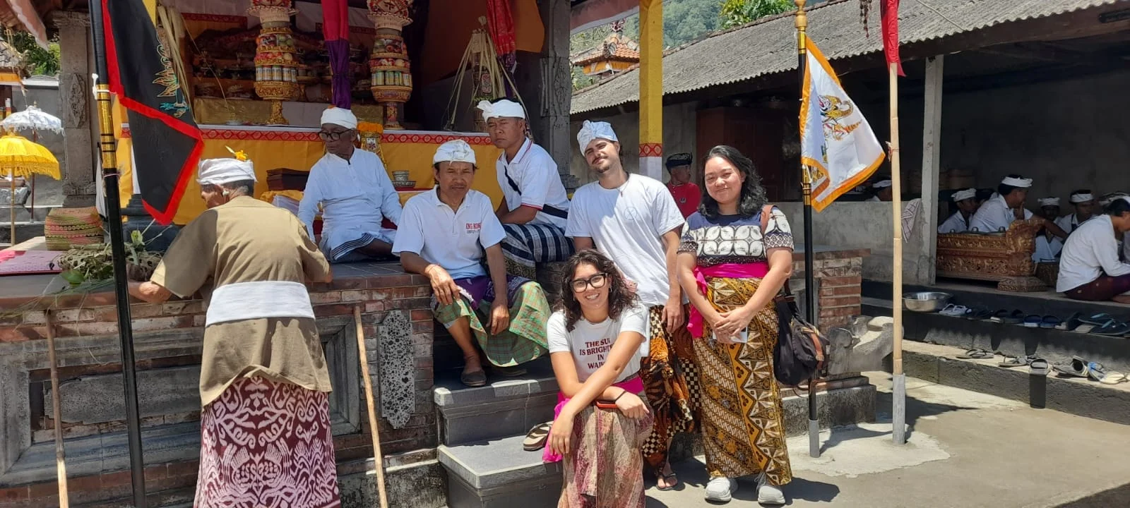 A group of volunteers and local people, some in traditional sarongs and white headwear, sitting and standing on the steps of a decorated temple courtyard.