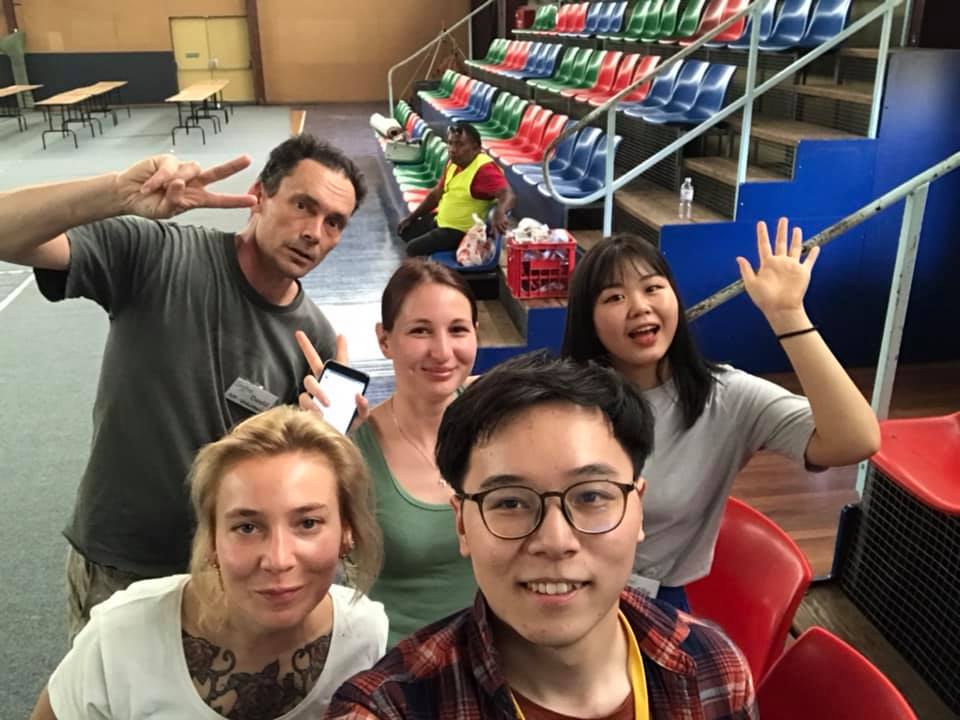 Five volunteers from different countries taking a selfie in a showground pavilion, smiling and waving, with another volunteer in a high-visibility vest behind them.