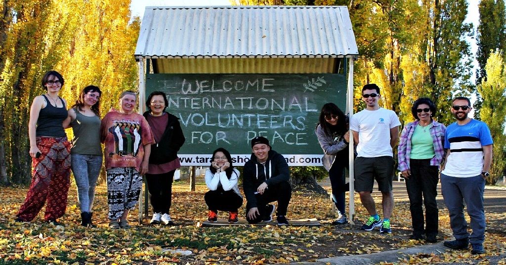 Ten volunteers standing and crouching around a chalkboard sign that reads “Welcome International Volunteers for Peace”, under yellow autumn trees.