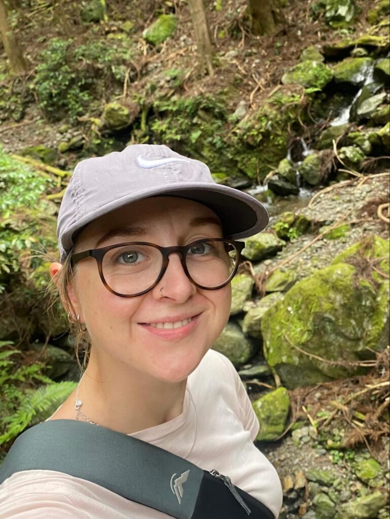 Charlotte smiling in a cap and glasses beside a mossy stream.