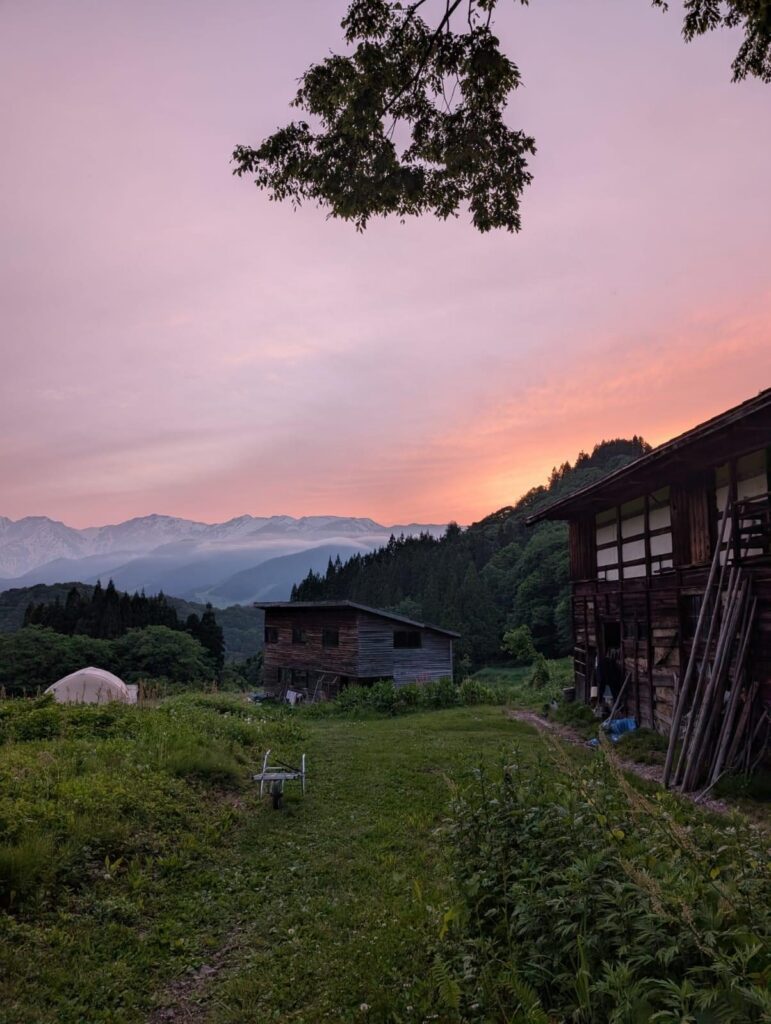 A pink sunset over distant mountains, with a wooden village house in the foreground.