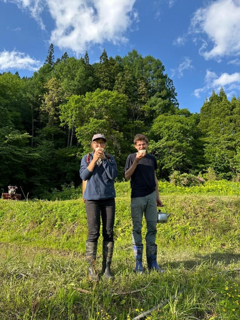 Two volunteers in gumboots standing in a sunny field, drinking tea.