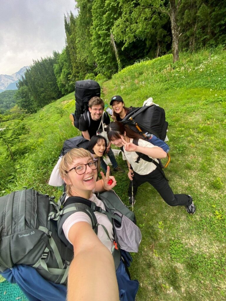 A selfie of five volunteers with large backpacks resting on a grassy hillside.